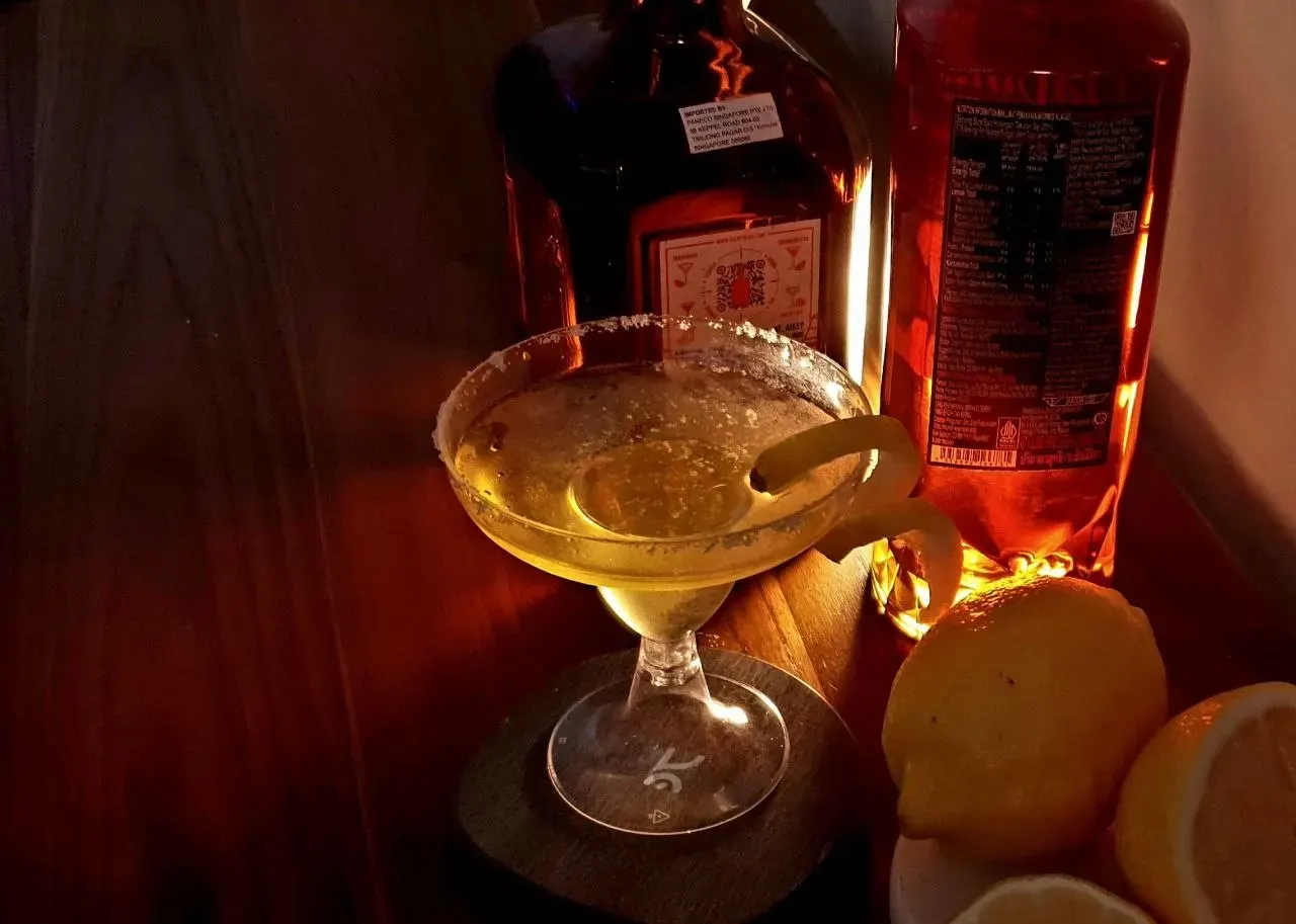 A coupe of a golden cocktail with a lemon twist, lit warm against dark wood